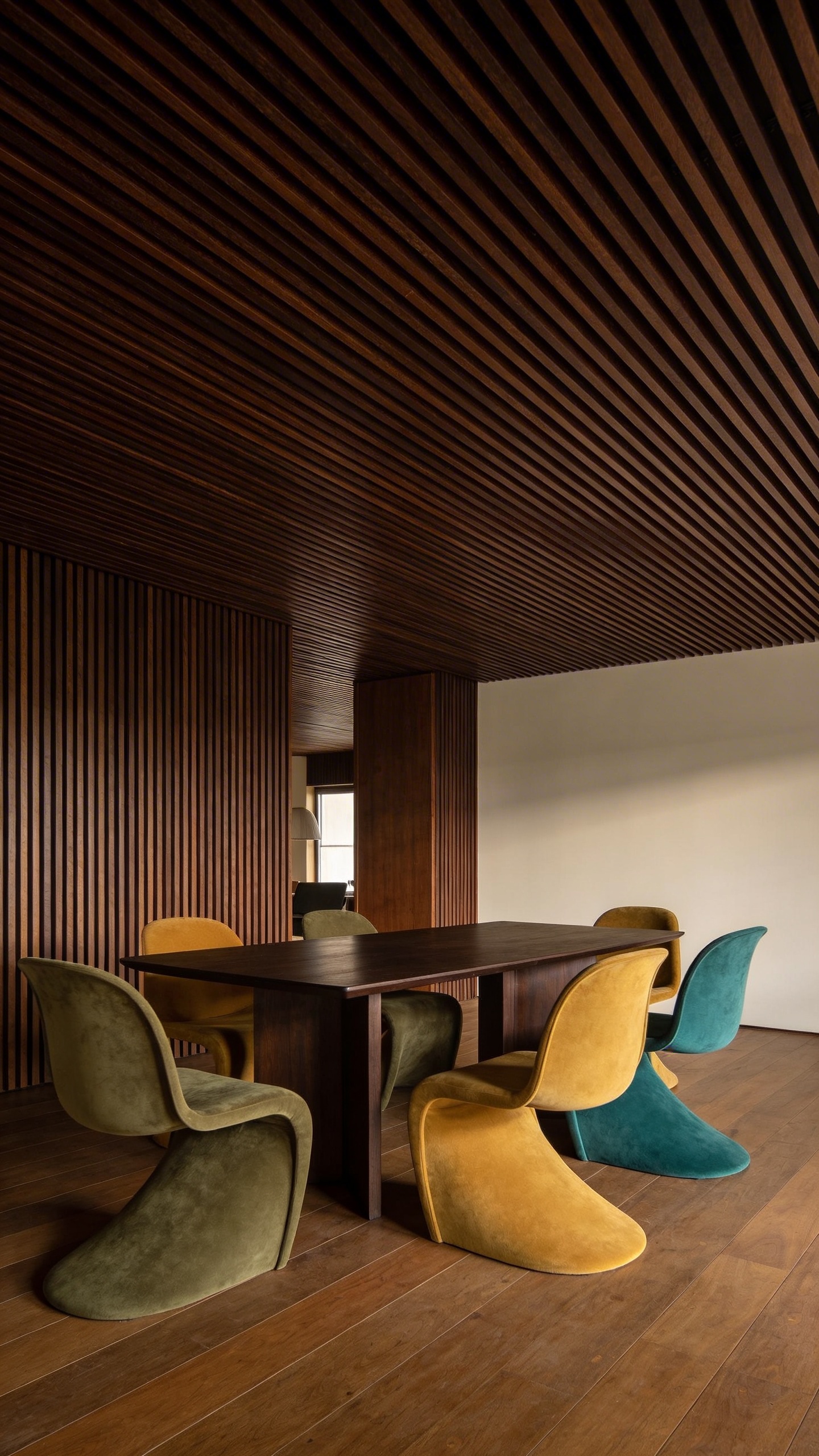 A high-quality, realistic photograph of a sophisticated mid-century modern dining room with a contemporary twist. The most striking feature is the dark wood slatted ceiling, composed of thin, evenly spaced dark walnut slats running parallel across the entire ceiling, providing rich texture and a subtle directional flow. The dining area features a sleek, dark wood dining table with clean lines, surrounded by organically shaped dining chairs upholstered in a muted color palette of olive green, mustard yellow, and teal. The room exudes an atmosphere of retro chic and sophisticated simplicity, bathed in soft, natural light that highlights the architectural details and the warm wood tones. No text.